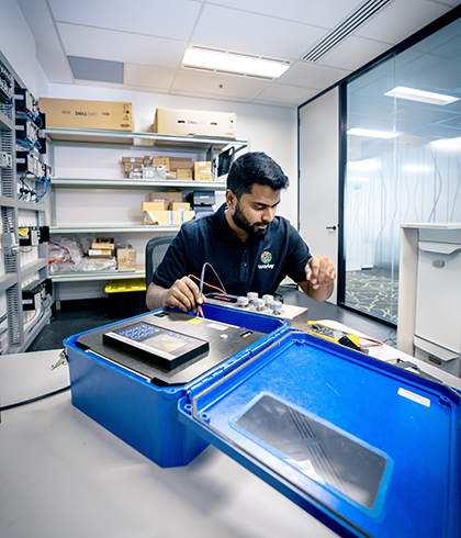 Optravation Energytechnician testing electronic components in a lab with tools, blue case, and shelves of equipment.